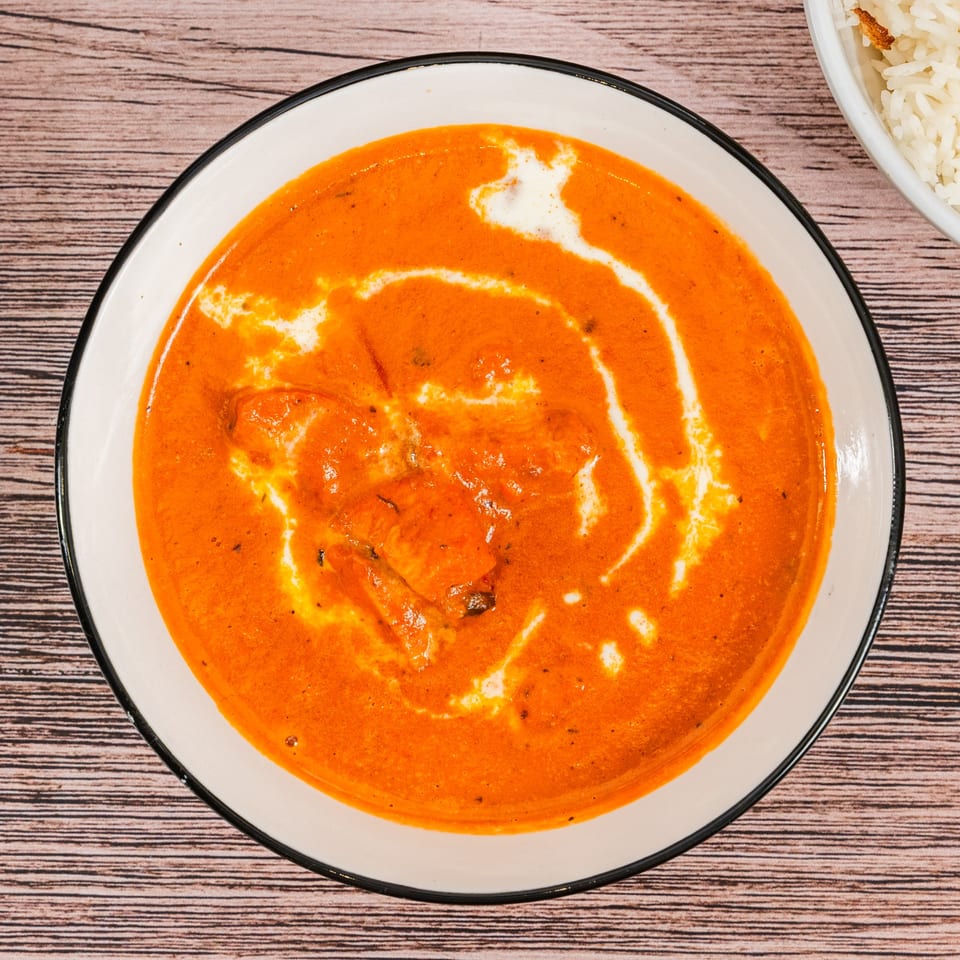 A bowl of Moh Moh Licious Indian butter chicken with a swirl of cream, next to rice.