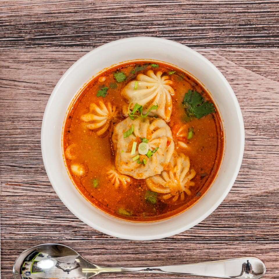 Moh Moh Licious Indian momo soup, a white bowl filled with dumplings in a rich orange broth.